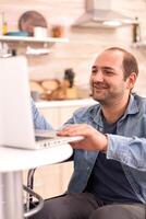 Guy in wheelchair smiling on video conference using laptop in kitchen. Disabled paralyzed handicapped man with walking disability integrating after an accident. photo