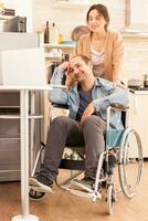 Positive man in wheelchair and wife looking at laptop in kitchen. Disabled paralyzed handicapped man with walking disability integrating after an accident. photo