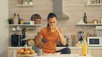 mujer Bebiendo Fresco naranja jugo en el Mañana mientras hojeada en teléfono inteligente en cocina. ama de casa utilizando moderno tecnología y Bebiendo saludable, natural, hecho en casa naranja jugo. refrescante Mañana foto