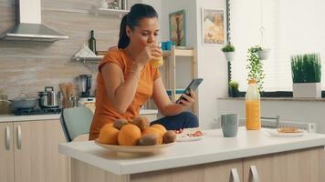 Bebiendo Fresco naranja jugo y hojeada en teléfono inteligente durante desayuno. ama de casa utilizando moderno tecnología y Bebiendo saludable, natural, hecho en casa naranja jugo. refrescante Mañana foto