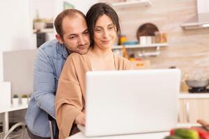 Smiling couple using laptop in kitchen. Man hugging wife. Happy loving cheerful romantic in love couple at home using modern wifi wireless internet technology photo