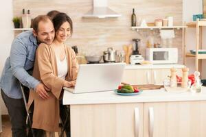 Cheerful couple using laptop in kitchen reading online recipe for breakfast. Happy loving cheerful romantic in love couple at home using modern wifi wireless internet technology photo