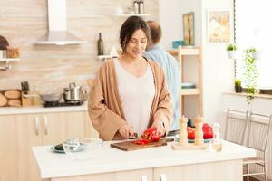 Novia rebanar rojo campana pimienta para ensalada en cocina. novio en el antecedentes. hermosa alegre contento sonriente mujer en brillante habitación preparando sano y orgánico comida. foto