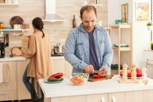Husband chopping tomatoes for salad on wooden cutting board in kitchen. Wife holding paper groceries bag. Happy in love cheerful and carefree couple helping each other to prepare meal photo