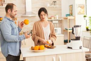 Couple preparing fresh smoothie in kitchen. Woman peeling off oranges. Husband doing tricks with fruit for wife. Healthy carefree and cheerful lifestyle, eating diet and preparing breakfast in cozy sunny morning photo