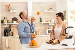 Man doing tricks with oranges for wife in kitchen while preparing healthy smoothie. Healthy carefree and cheerful lifestyle, eating diet and preparing breakfast in cozy sunny morning photo