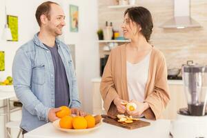 Couple smiling at each other while preparing nutritious smoothie. Woman peeling off orange. Healthy carefree and cheerful lifestyle, eating diet and preparing breakfast in cozy sunny morning photo