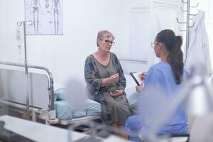 Nurse taking notes on clipboard consulting old woman, asking questions to mature patients filling disease history form, prescribing treatment. Senior old woman talking with young assistent, healthcare concept photo