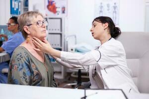 joven médico palpando cuello de mayor mujer, mayor paciente visitando médico a hospital comprobación tiroides garganta conmovedor salud a clínica. cuidado de la salud especialista, seguro médico del estado, tratamiento médico concepto. foto