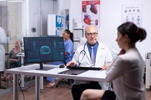 mayor médico explicando cuerpo escanear a joven dama desde computadora, hembra paciente visitando médico narración acerca de salud quejas, médico facultativo preguntando preguntas hablando acerca de relleno enfermedad. foto