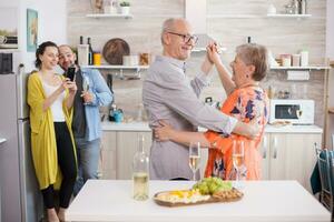 Happy senior couple dancing in kitchen during family celebration. Delicious assorted cheese on wooden board. Woman taking photos. photo