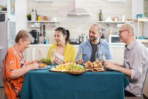 Senior woman laughing while having lunch with family in kitchen. Cheerful senior father holding wine glass. Tasty potatoes. photo