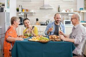 Father and son holding wine glasses during family brunch. Daughter and senior mother looking at smartphone. Tasty seasoned potatoes. photo