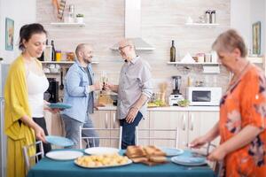 Father and son toasting with wine and smiling at each other. Mother and daughter setting plates on table for lunch. photo