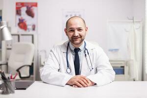 retrato de joven médico sonriente mirando a el cámara en hospital oficina vistiendo blanco Saco y estetoscopio. foto