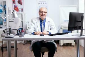 Senior doctor smiling at camera sitting in medical office during video meeting, Professional man wearing white coat talking by conference call chat consulting patient online looking at webcam photo