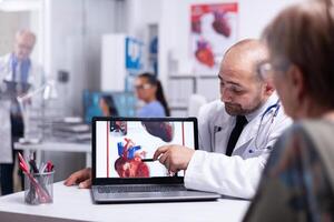 Physician using laptop showing heart disease to elderly woman pointing on laptop sitting in hospital room talking with senior patient. Team of doctors working in background using clipboard and xray photo