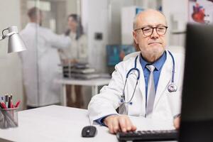 Close up of physician with grey hair typing document about new drug in hospital cabinet and young medic discussing with patient in the background wearing white coat. photo