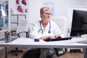 mayor hembra médico en laboratorio abrigo, lentes y estetoscopio terminado su cuello mecanografía en computadora sentado en médico oficina utilizando mirando a escritorio sonriente mientras trabajando en médico oficina foto