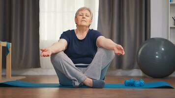 Senior woman meditating sitting on yoga mat in living room. Active healthy lifestyle sporty old person training workout home wellness and indoor exercising photo