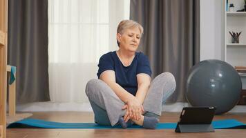 Senior woman warming up shoulder watching online tutorial sitting on yoga mat. Online learning and study, Active healthy lifestyle sporty old person training workout home wellness and indoor exercising photo