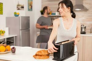 Woman using toaster to roast bread in kitchen during breakfast. Young housewife at home cooking morning meal, cheerful with affection and love photo