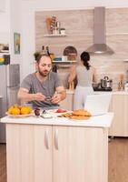 Independence worker eating breakfast and working from home while his wife cooks. Freelancer working remote, talking in videoconference video call online web internet meeting from home, communication device photo