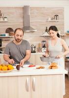 esposa Cocinando huevos para marido durante desayuno mientras él es manchando mantequilla en asado un pan. vistiendo pijama en el mañana, preparando comida juntos, joven contento Pareja amor y matrimonio foto