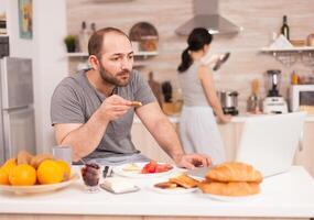 Entrepreneur on a video conference while eating breakfast in kitchen. Freelancer working remote, talking in videoconference video call online web internet meeting from home, communication device photo