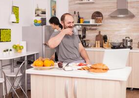 Entrepreneur working from home while eating breakfast in kitchen, wearing pajamas enjoying roasted bread with butter. Freelancer working online via internet using modern digital technology photo