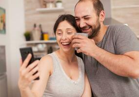 Happy young couple taking selfies during breakfast. Joyful married husband and wife making funny faces while taking a photo during breakfast in kitchen.