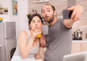 gracioso Pareja tomando selfies durante desayuno. alegre casado marido y esposa haciendo gracioso caras mientras tomando un foto durante desayuno en cocina.