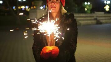 Lycklig leende kvinna i en röd hatt står på de gata, innehav sparklers i henne händer. bengal ljus, festlig feyerferk. vinter- roligt på de gata jul eller ny år firande. långsam rörelse video