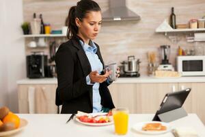 Young business woman in the kitchen having a healthy meal while talking on a video call with her colleagues from the office, using modern technology and working around the clock photo