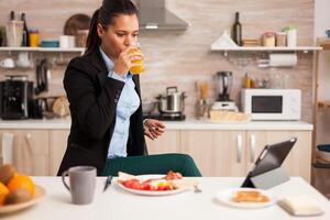 Businesswoman drinking fresh juice during breakfast before work watching a video on tablet. Business woman reading the last news online before going to work, using modern technology in the kitchen while eating a healthy meal photo