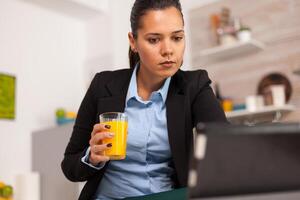 Businesswoman checking email at home in the kitchen drinking an orange juice. Business woman reading the last news online before going to work, using modern technology in the kitchen while eating a healthy meal photo