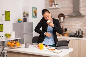 Business woman eating toasted bread with butter while working on laptop during breakfast. Concentrated business woman in the morning multitasking in the kitchen before going to the office, stressful way of life, career and goals to meet photo