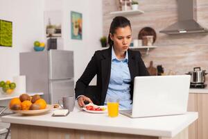 Business woman eating breakfast and working on laptop. Concentrated business woman in the morning multitasking in the kitchen before going to the office, stressful way of life, career and goals to meet photo
