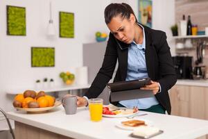 Business woman reading a bad news on her tablet while eating breakfast. Concentrated business woman in the morning multitasking in the kitchen before going to the office, stressful way of life, career and goals to meet photo