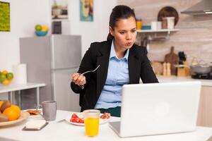 Businessperson eating breakfast while working from home. Concentrated business woman in the morning multitasking in the kitchen before going to the office, stressful way of life, career and goals to meet. photo