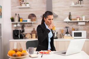 Young business woman enjoying the breakfast and looking at laptop. Business woman reading the last news online before going to work, using modern technology in the kitchen while eating a healthy meal photo