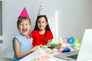 niños en línea cumpleaños fiesta. pequeño muchachas en vestidos, sombrero celebrar fiesta con amigos. conferencia, vídeo llamada en computadora portátil, computadora. cuarentena, coronavirus pandemia COVID-19. foto