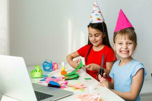 niños en línea cumpleaños fiesta. pequeño muchachas en vestidos, sombrero celebrar fiesta con amigos. conferencia, vídeo llamada en computadora portátil, computadora. cuarentena, coronavirus pandemia COVID-19. foto