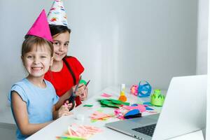 niños en línea cumpleaños fiesta. pequeño muchachas en vestidos, sombrero celebrar fiesta con amigos. conferencia, vídeo llamada en computadora portátil, computadora. cuarentena, coronavirus pandemia COVID-19. foto