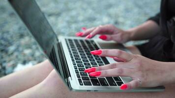 geslaagd bedrijf vrouw met helder rood manicure typen Aan laptop toetsenbord buitenshuis Aan strand met zee visie. dichtbij omhoog vrouw handen schrijven Aan computer. freelance, digitaal nomade, reizen en vakantie. video