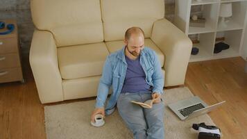 hombre en mezclilla camisa riendo mientras leyendo un libro y Bebiendo algunos café en el piso foto