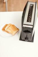 Roasted bread near electric toaster on the table in the kitchen. Housewife using bread toaster for delicious breakfast. Healthy morning in cozy interior, delicious home meal preparation. photo