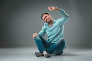 Handsome young man sitting on a floor with raised hands, isolated on gray background photo