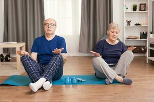 Senior couple relaxing sitting on yoga mat in the middle of the room. Old person healthy lifestyle exercise at home, workout and training, sport activity at home on yoga mat. photo