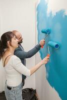 Happy couple giving the wall a makeover with blue paint using roller brushes. Home during renovation, decoration and painting. Interior apartment improvement maintenance. Roller, ladder for house repair photo